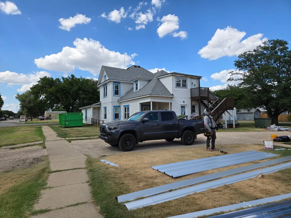 Metal roofing panels staged in front of a historic farmhouse mid re-roof, with dumpster and work truck on site